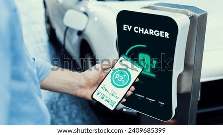 Businessman holding smartphone display battery status interface by smart EV mobile application while EV car recharging electricity from charging station in car park. Peruse