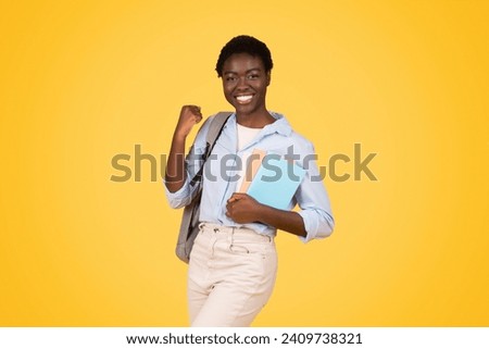 Positive teen african american lady student with books, exuding triumph and joy, raises fist in victorious gesture, isolated on yellow background, studio. Celebrate success and win, study