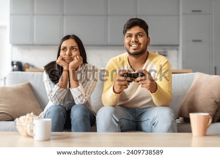 Bored young indian woman sitting near boyfriend playing video games at home, unhappy eastern female feeling lonely, cheerful man holding joystick and ignoring his upset girlfriend, free space