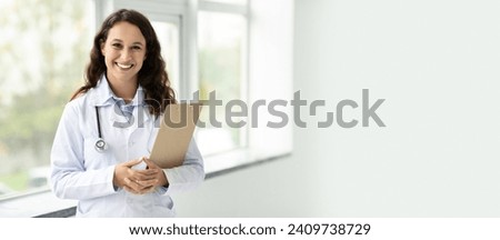Smiling young brunette european woman doctor therapist in white coat holding clipboard, posing next to window at modern clinic, copy space, web-banner. Medicine service, health care and treatment