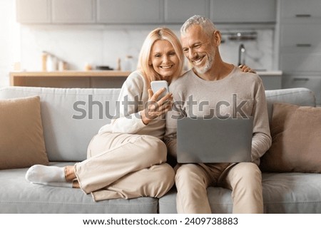 Retired european couple spending time at home, using gadgets, Happy senior man and woman spouses sitting on couch, using laptop and smartphone, checking photos, websurfing, copy space Royalty-Free Stock Photo #2409738883
