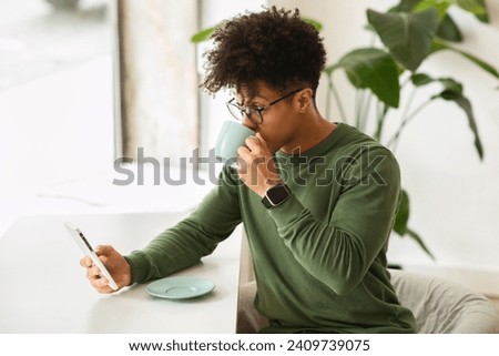 Relaxed stylish millennial black man wearing eyeglasses drinking coffee and using smartphone at cafe, texting friends, scrolling, websurfing, looking through window, copy space