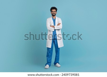 Smiling young arab doctor man posing with folded arms over blue studio background, confident middle eastern male physician wearing uniform and stethoscope, full body length, copy space