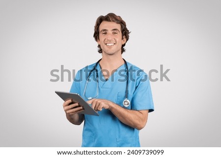 Happy young male nurse wearing blue scrubs and a stethoscope using a digital tablet, possibly for patient records or telehealth services Royalty-Free Stock Photo #2409739099