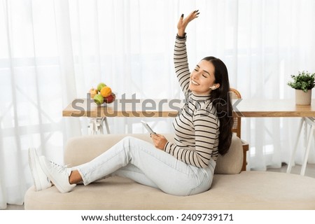 Happy millennial arab woman with smartphone in wireless headphones sit at sofa, listen music, enjoy spare time, dance in living room interior. Spare time and fun, audio app, rest at home Royalty-Free Stock Photo #2409739171