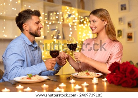 Smiling european couple engaging in toast with red wine glasses over candlelit dinner table, with a backdrop of twinkling fairy lights and roses Royalty-Free Stock Photo #2409739185