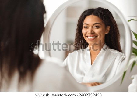 Attractive Young Black Woman Wearing White Silk Robe Looking In Mirror At Home, Beautiful African American Lady Enjoying Her Reflection, Making Skincare And Beauty Routine, Selective Focus Royalty-Free Stock Photo #2409739433