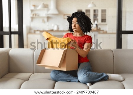 Surprised young black woman opening delivery box and looking at yellow t-shirt, shocked african american lady reacting to an unexpected item in parcel, sitting on a sofa in bright, modern living room Royalty-Free Stock Photo #2409739487