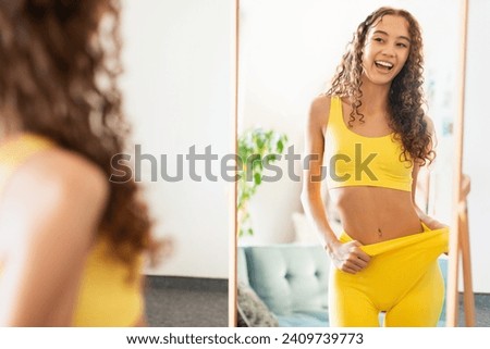 Sporty caucasian teenager girl pulling fitness pants waist, showing slimming result smiling at her mirror reflection, admiring slender shape, motivated after workout session at home Royalty-Free Stock Photo #2409739773