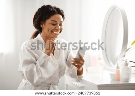 Beautiful smiling black woman using smartphone while sitting at dressing table at home, smiling african american lady wearing white silk robe reading beauty blog or watching video online, copy space Royalty-Free Stock Photo #2409739861