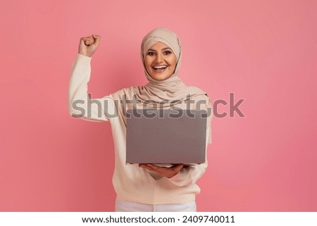 Joyful muslim woman in hijab holding laptop and shaking her fist, symbolizing her success and excitement, standing against pink background, epitomizing achievement in digital age, copy space