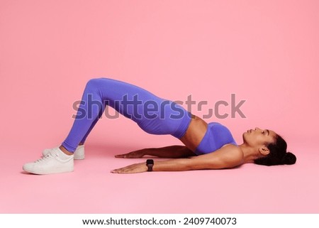 Young black woman in activewear practicing yoga bridge pose on studio floor over pink backdrop, embodying strength and wellness. Side view, full length shot. Fitness workout