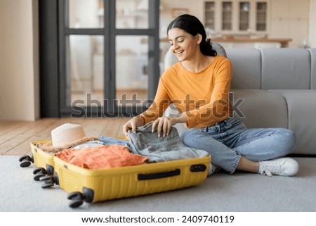 Smiling young indian woman sitting on the floor, contentedly packing clothes into her open yellow suitcase, happy eastern female getting ready for trip, preparing luggage for journey, copy space Royalty-Free Stock Photo #2409740119