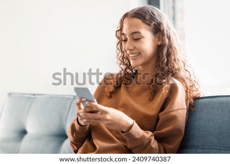 European adolescent girl browsing internet on phone, involved in online communication and application use, sitting carefree and happy in her cozy home setting. Mobile apps, gadgets
