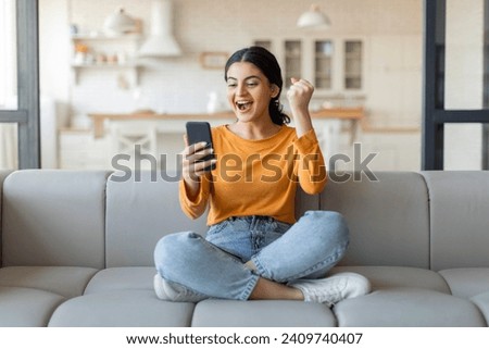 Joyful young indian woman with smartphone celebrating victory. happy excited female sitting on couch at home, holding mobile phone and shaking fist, cheering success and online win, got good news Royalty-Free Stock Photo #2409740407