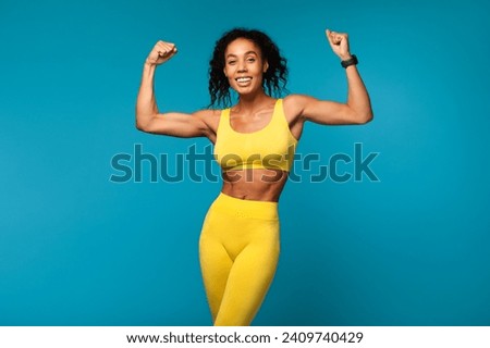 Strength And Power. Happy young sporty black lady in sportswear celebrating win and success, shaking and raising clenched fists, showing her biceps arm muscles over blue studio background