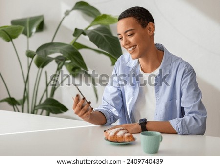 Content young latin lady enjoys moment of leisure, smiling, reading message on smartphone, with croissant and coffee cup on desk. Modern relaxation and communication, enjoy snack and chat