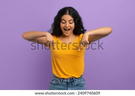 Excited amazed young indian woman with curly hair pointing down and grimacing, showing great offer, deal, advertisement, isolated on purple studio background