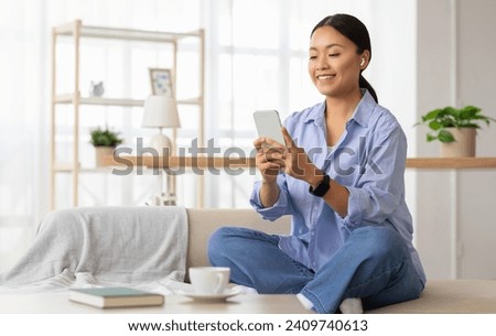 Cheerful millennial asian woman in casual clothing sitting on couch in cozy living room interior, using smartphone and wireless earphones at home, drinking coffee, watching podcast, video, copy space