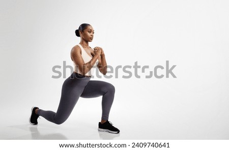 African American fitness woman stretching legs performing forward lunges over white studio backdrop, enjoying workout routine, looking aside. Full length shot, empty space Royalty-Free Stock Photo #2409740641