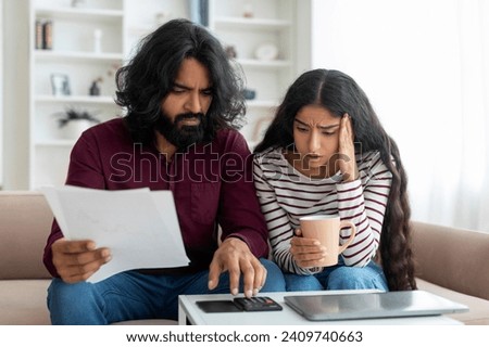 Shocked Married Young Indian Couple Planning Budget Together, Reading Papers And Calculating Spends While Sitting On Couch In Living Room, Husband And Wife Checking Documents And Accounting Taxes Royalty-Free Stock Photo #2409740663