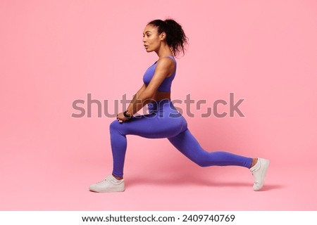 Young black woman in activewear performing forward lunges on pink studio backdrop, displaying fitness routine, making stretching legs exercise looking aside, side view shot