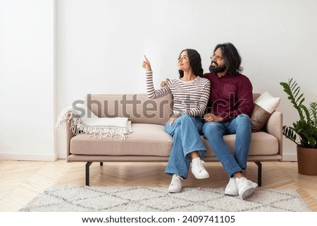 Lovely smiling young indian couple wearing casual outfit sitting on couch at home, brunette woman pointing at copy space, showing her husband nice offer, living room interior. Mortgage, moving Royalty-Free Stock Photo #2409741105