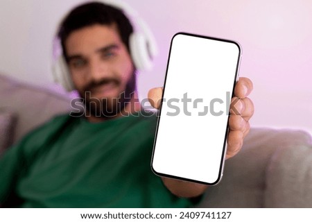 Cheerful bearded man in a green t-shirt enjoying music through white headphones, presenting a smartphone with a blank screen, with a soft-focus background. App blog, website