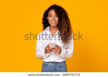 Happy african american woman holds her mobile phone, websurfing and using applications looking at camera with smile, over yellow studio background. Modern gadgets and communication Royalty-Free Stock Photo #2409741715