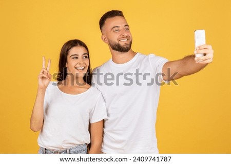 A beaming glad caucasian young woman flashes a peace sign while taking a selfie with a smiling man, capturing a moment of joy and friendship on a sunny yellow background, studio