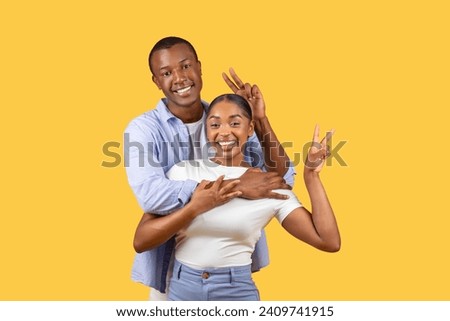 Black man and woman in joyful embrace make peace signs with their hands, smiling brightly, communicating vibe of fun and positivity on yellow backdrop