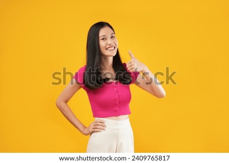 Photo of young asian woman ethnicity 20s wearing pink t-shirt showing thumb up like gesture isolated on pastel plain yellow color background studio portrait. People lifestyle concept