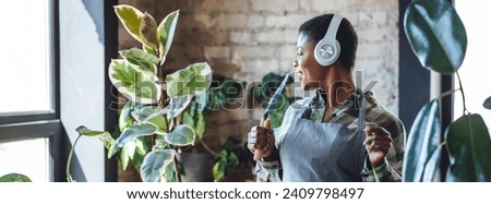 Concept of wellbeing, relaxation, work life balance, simple pleasures. Beautiful smiling plus size African American woman is doing home gardening, repotting, taking care about plants. Banner Royalty-Free Stock Photo #2409798497