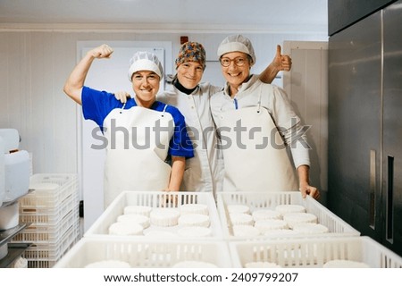 Smiling female dairy factory worker showing thumb up while embracing colleague demonstrating bicep standing near ready ricotta cheese after successful work