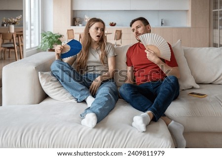 Family wife husband suffering from stuffiness waving blue paper fans at home. Tired exhausted man woman feel bad in hot weather sit on couch in room without air conditioner. Overheating, sultriness. Royalty-Free Stock Photo #2409811979
