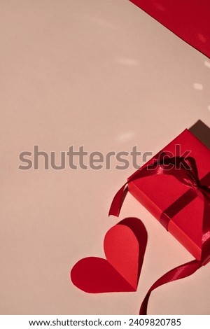 Red gift box and paper heart shaped valentine card on peach beige background with bright light and harsh shadows. Minimal Valentine day celebration concept, business brand template.