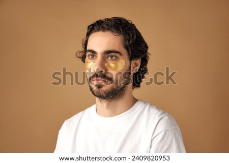 Portrait of calm brunette man with beard applying hydrogel patches with moisturized effect under eyes against sandy color studio background. Concept of facial care routine, cosmetic product.
