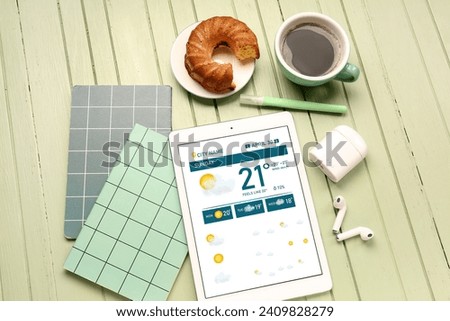 Tablet computer with weather forecast, notebooks, earphones and breakfast on green wooden background