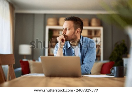 A focused, deep in thought male freelance copywriter looking aside with his hand on his chin sitting in front of a laptop having a writer's block. Royalty-Free Stock Photo #2409829599