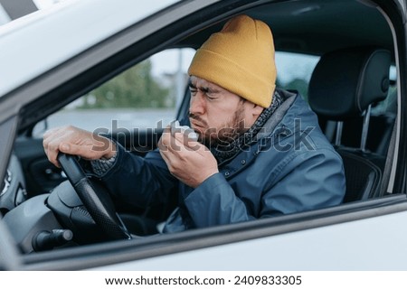 Join a young chauffeur on a unique journey through the challenges of flu season. This 4k image encapsulates the bearded man's discomfort, showcasing sneezing and coughing on city streets. Royalty-Free Stock Photo #2409833305
