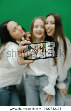 three beautiful young girls in a beauty salon on a green background look at the camera, take a selfie on the phone and smile, happy girlfriends in jeans and white shirts