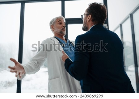 Violence against health workers. Patient is aggressive, have conflict with doctor in hospital corridor. Visitor holding doctor by coat, screaming, threatening him. Royalty-Free Stock Photo #2409869299