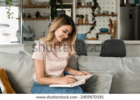 Young woman sitting on the sofa writing down the guest list of her friends whom she will invite to her bachelorette party for her wedding. Creative girl writes down ideas make a list of things to do Royalty-Free Stock Photo #2409870181