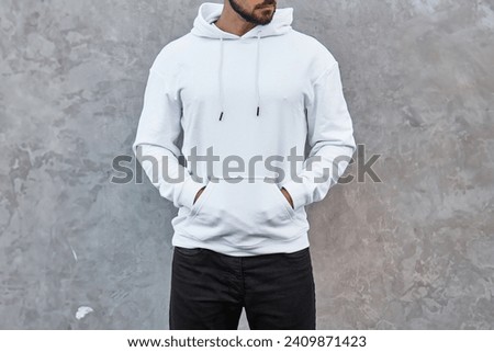 A mock-up template of a white hoodie worn by a man. A design concept for print and branding. A young man poses outdoors in a casual and trendy streetwear style. No face is shown.