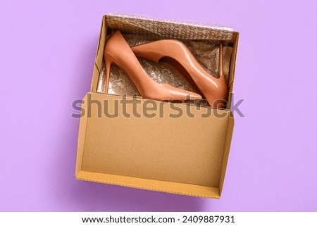 Cardboard box with high heeled shoes on lilac background