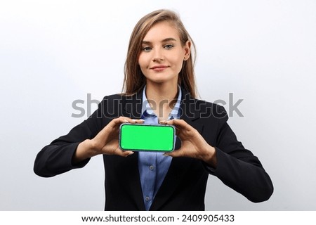 Caucasian fair-haired young woman in a jacket shows a green screen of a smartphone for advertising.