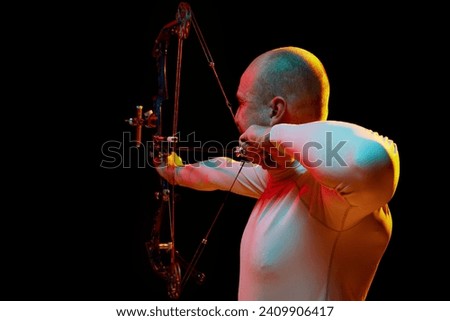 Back view image of man, archery at hele aiming with archery bow on target against black studio background in neon light. Concept of professional sport and hobby, competition, action, game