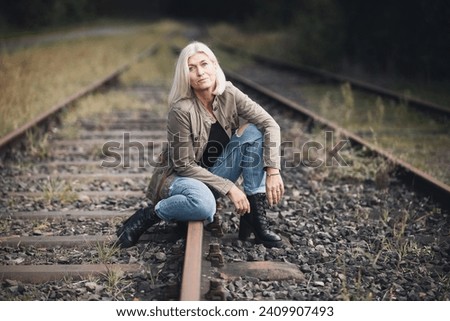 An older woman in her fifties with blonde hair, dressed in a brown jacket and jeans, sits with her legs bent on a railway track. Royalty-Free Stock Photo #2409907493