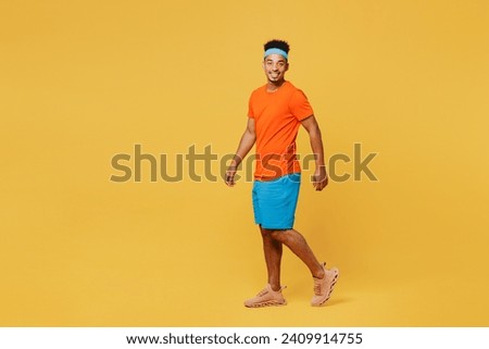 Full body side view happy young fitness trainer sporty man sportsman wear orange t-shirt walk go look camera spend time in home gym isolated on plain yellow background. Workout sport fit abs concept Royalty-Free Stock Photo #2409914755