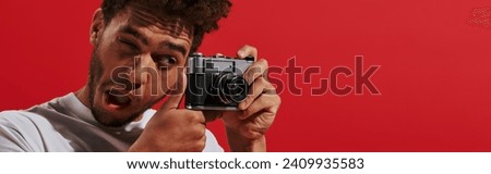 funny african american man looking at viewfinder while taking shot on red background, banner